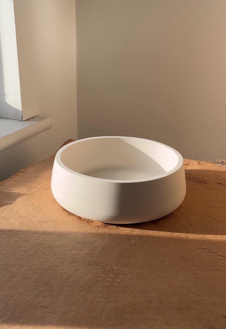May include: A round, off-white bowl with a wide, gently sloping rim. The bowl is made of a solid material and sits on a wooden surface. The bowl's interior is visible, and the lighting creates shadows.