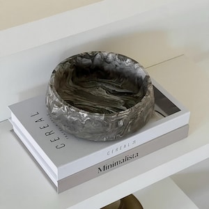 May include: A round, marbled gray and white decorative bowl sits atop two books on a white shelf. The books are titled "Cereal" and "Minimalista". The bowl has a swirling pattern and a smooth finish.