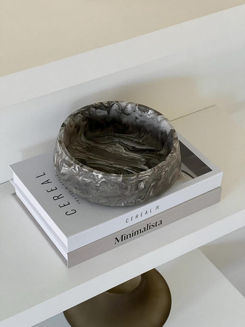 May include: A round, marbled gray and white decorative bowl sits atop two books on a white shelf. The books are titled "Cereal" and "Minimalista". The bowl has a swirling pattern and a smooth finish.