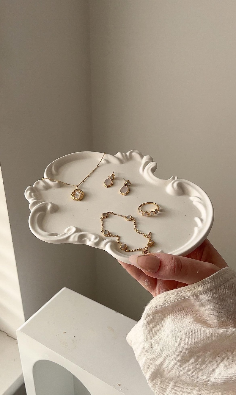 May include: A decorative white tray displays gold-coloured jewellery. The jewellery includes a necklace, earrings, a bracelet, and a ring, all with intricate designs and small, round, light-coloured stones. The tray is held by a person.