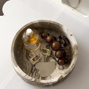 May include: A round, marbled decorative tray holds various accessories. Items include a perfume bottle, gold hoop earrings, brown hair clips, and a silver gua sha tool. A white shelf with a bracelet is in the background.