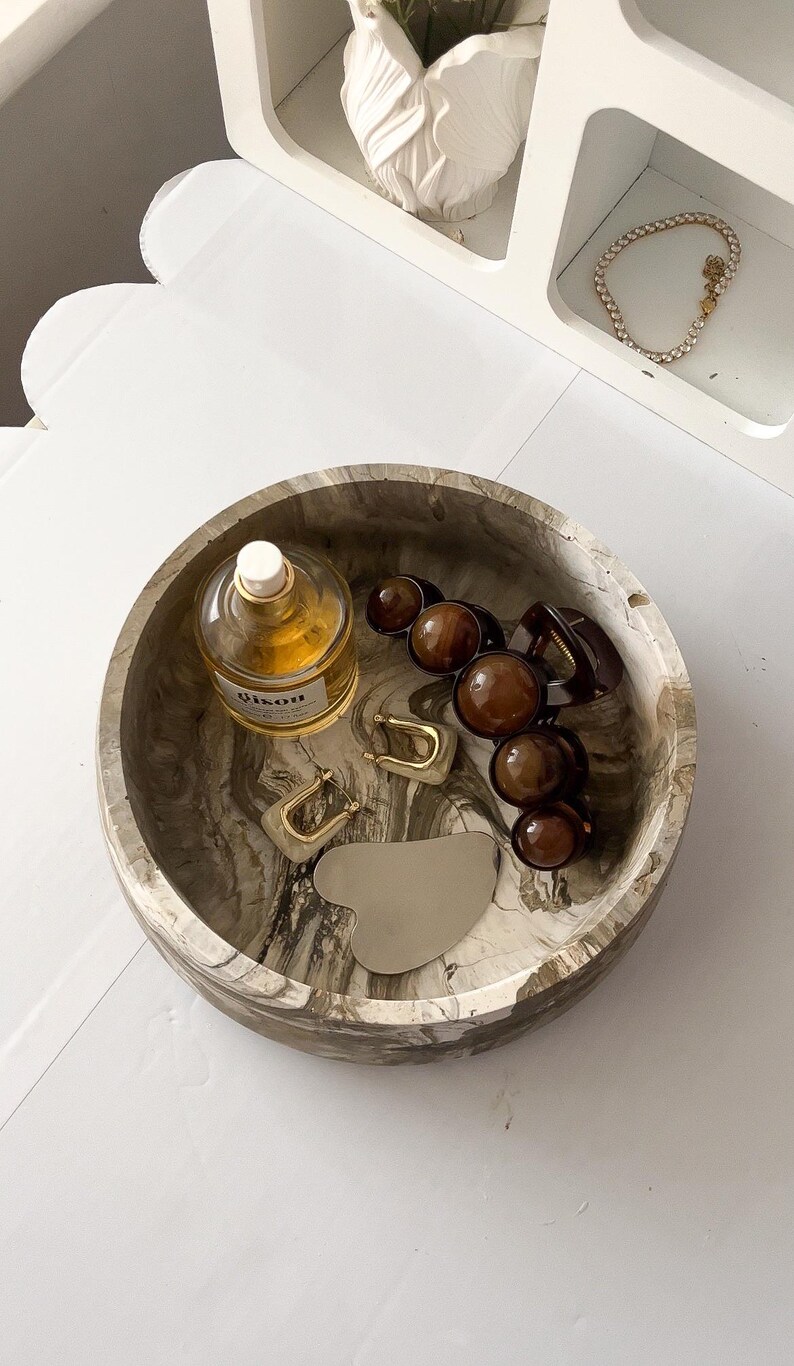 May include: A round, marbled decorative tray holds various accessories. Items include a perfume bottle, gold hoop earrings, brown hair clips, and a silver gua sha tool. A white shelf with a bracelet is in the background.