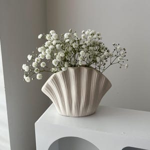 May include: A white ceramic vase shaped like a seashell with a bouquet of white baby's breath flowers.