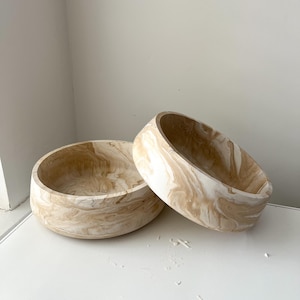 May include: Two decorative wooden bowls with a marbled beige and white pattern. The bowls are round with a wide opening and a thick, rounded base. One bowl is upright, and the other is tilted.