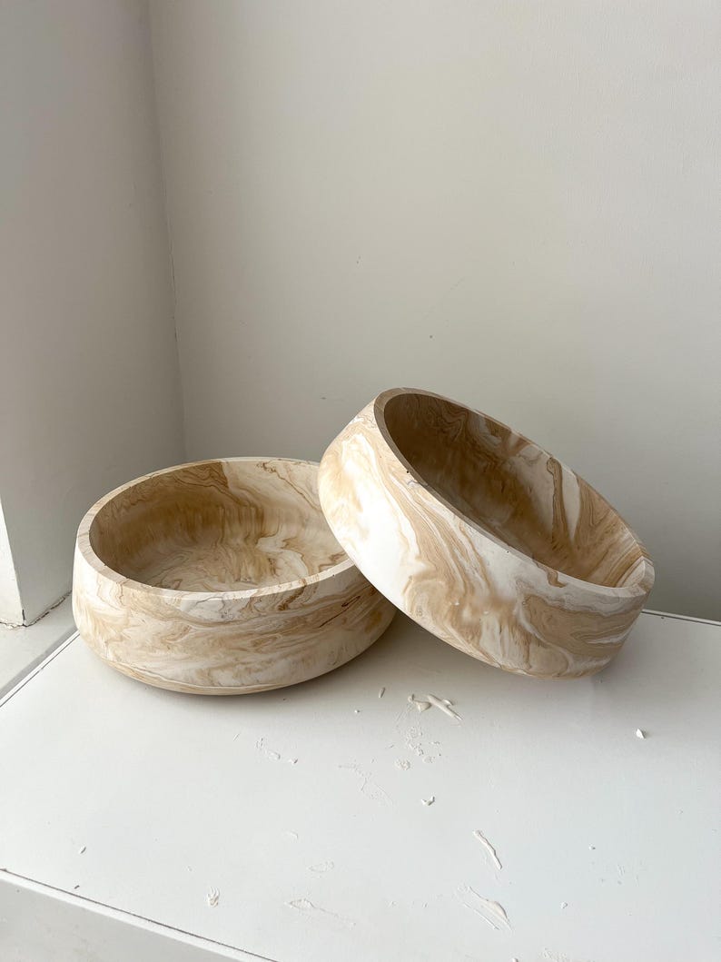 May include: Two decorative wooden bowls with a marbled beige and white pattern. The bowls are round with a wide opening and a thick, rounded base. One bowl is upright, and the other is tilted.