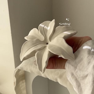 May include: A matte off-white ceramic flower sculpture with six distinct petals and a small circular opening in the center. The object features the word Neutral printed in white text above the petals, accompanied by two small four-pointed star icons.