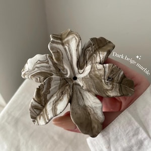May include: A flower-shaped object with a marbled pattern in shades of white and dark beige. The piece features five distinct petals with wavy edges and a small circular hole in the center. The text "Dark beige marble" is printed on the right side.