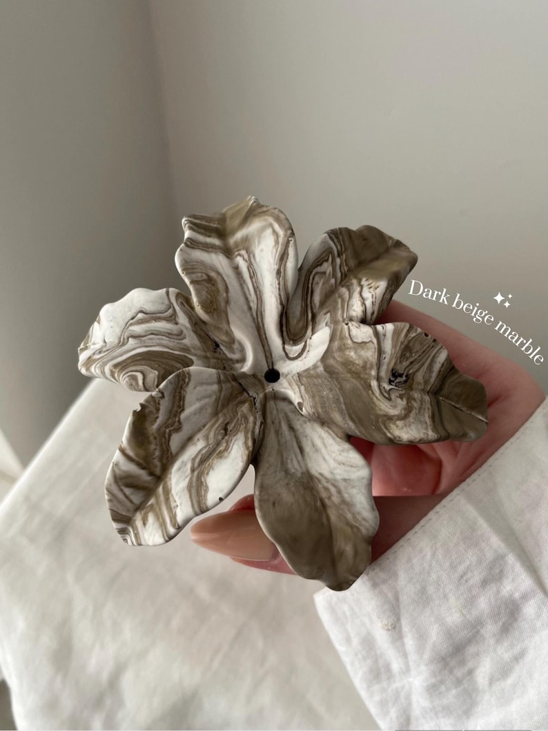 May include: A flower-shaped object with a marbled pattern in shades of white and dark beige. The piece features five distinct petals with wavy edges and a small circular hole in the center. The text "Dark beige marble" is printed on the right side.