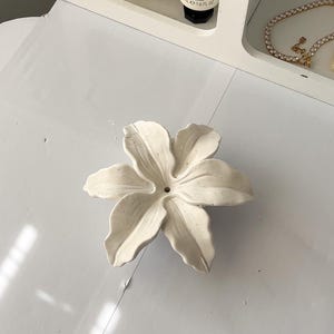 May include: A white ceramic flower-shaped dish with six curved petals and a small central hole. The surface has a matte, slightly textured finish with subtle speckling throughout the material, creating a natural appearance for this decorative item.
