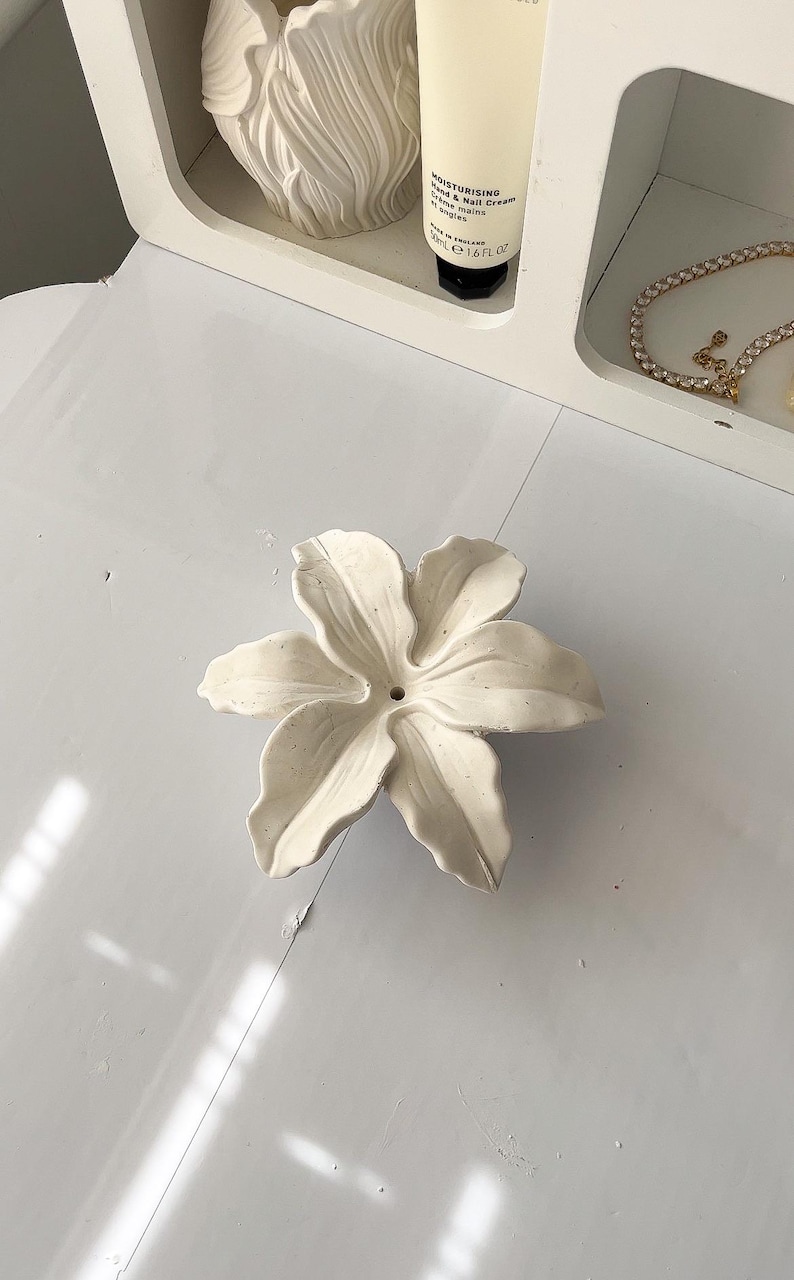 May include: A white ceramic flower-shaped dish with six curved petals and a small central hole. The surface has a matte, slightly textured finish with subtle speckling throughout the material, creating a natural appearance for this decorative item.