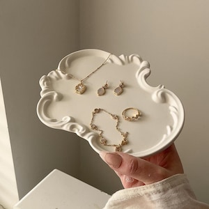 May include: A selection of gold-coloured jewellery, including a necklace, earrings, bracelet, and ring, presented on a white, cloud-shaped decorative tray. The jewellery features delicate designs and gemstones. The tray is held by a person.