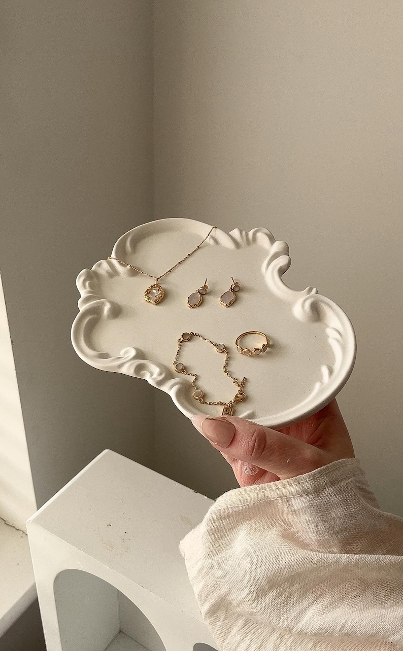 May include: A selection of gold-coloured jewellery, including a necklace, earrings, bracelet, and ring, presented on a white, cloud-shaped decorative tray. The jewellery features delicate designs and gemstones. The tray is held by a person.