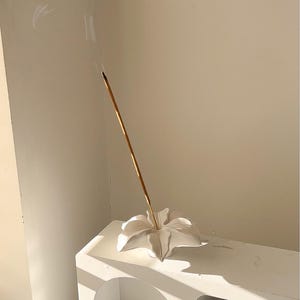 May include: A white ceramic incense holder shaped like a flower with a single lit incense stick emitting a thin trail of smoke. The holder is positioned on a white arched shelf unit that also contains a glass jar with a wooden lid and a hair clip.