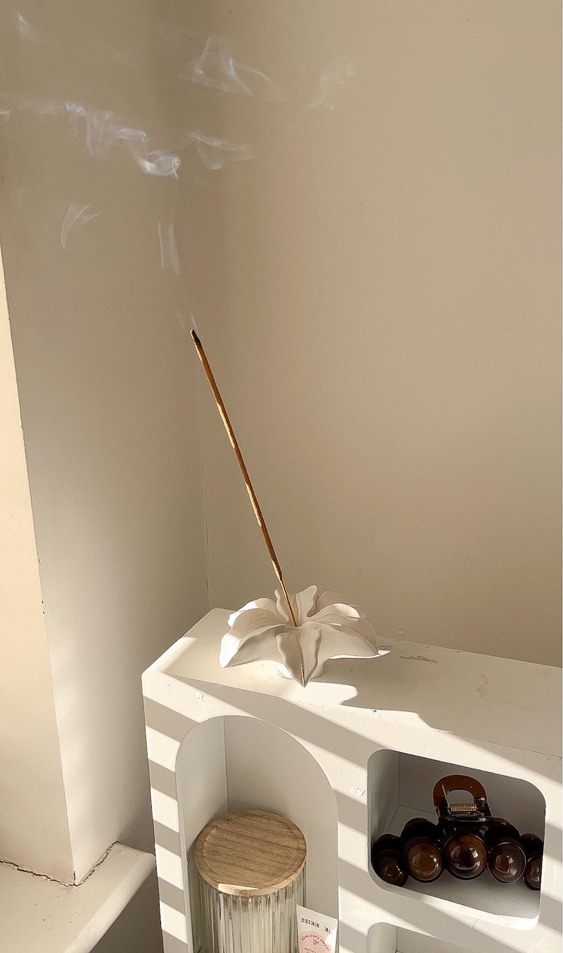 May include: A white ceramic incense holder shaped like a flower with a single lit incense stick emitting a thin trail of smoke. The holder is positioned on a white arched shelf unit that also contains a glass jar with a wooden lid and a hair clip.