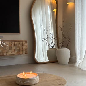 May include: A wooden coffee table with a lit candle and a large, irregularly shaped mirror. The candle is in a round, textured container. The mirror has a light wood frame. Two vases with branches are next to the mirror.