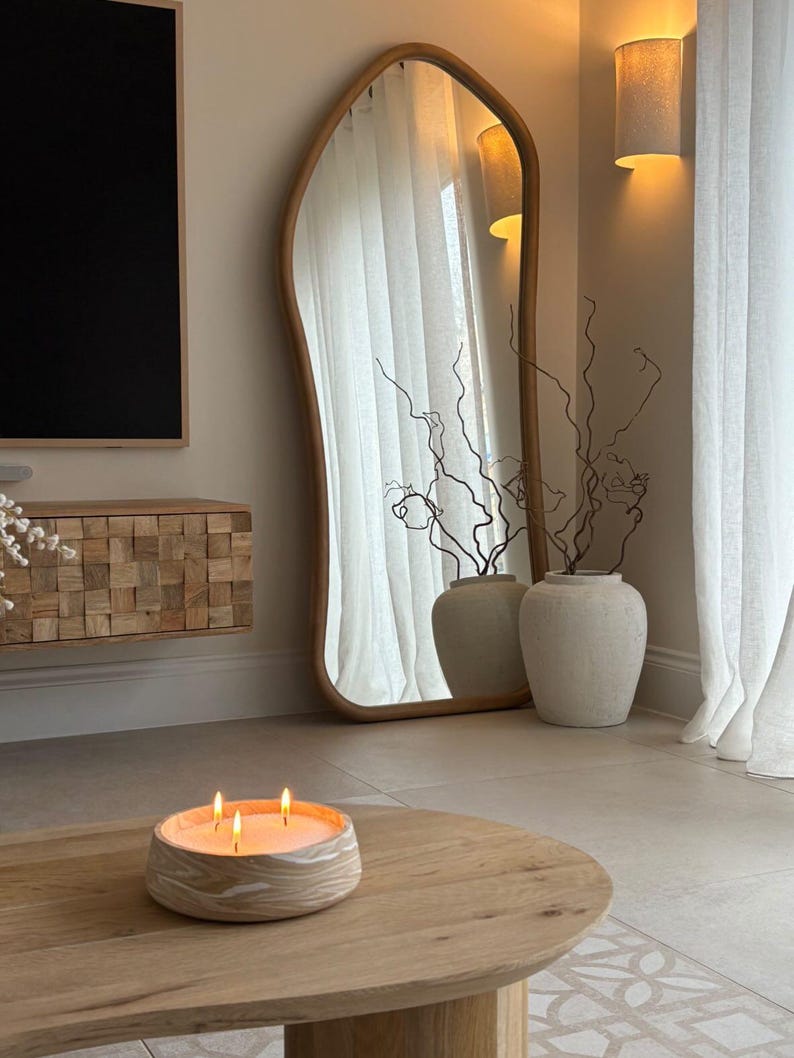 May include: A wooden coffee table with a lit candle and a large, irregularly shaped mirror. The candle is in a round, textured container. The mirror has a light wood frame. Two vases with branches are next to the mirror.