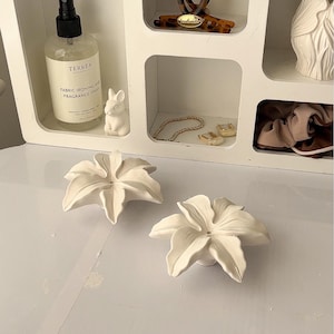 May include: Two white ceramic flower-shaped objects sit in the foreground. Behind them, a white shelving unit contains a clear spray bottle labeled Terrea Fabric Ironing and Fragrance Spray, a tortoiseshell hair clip, a white figurine, and jewelry.
