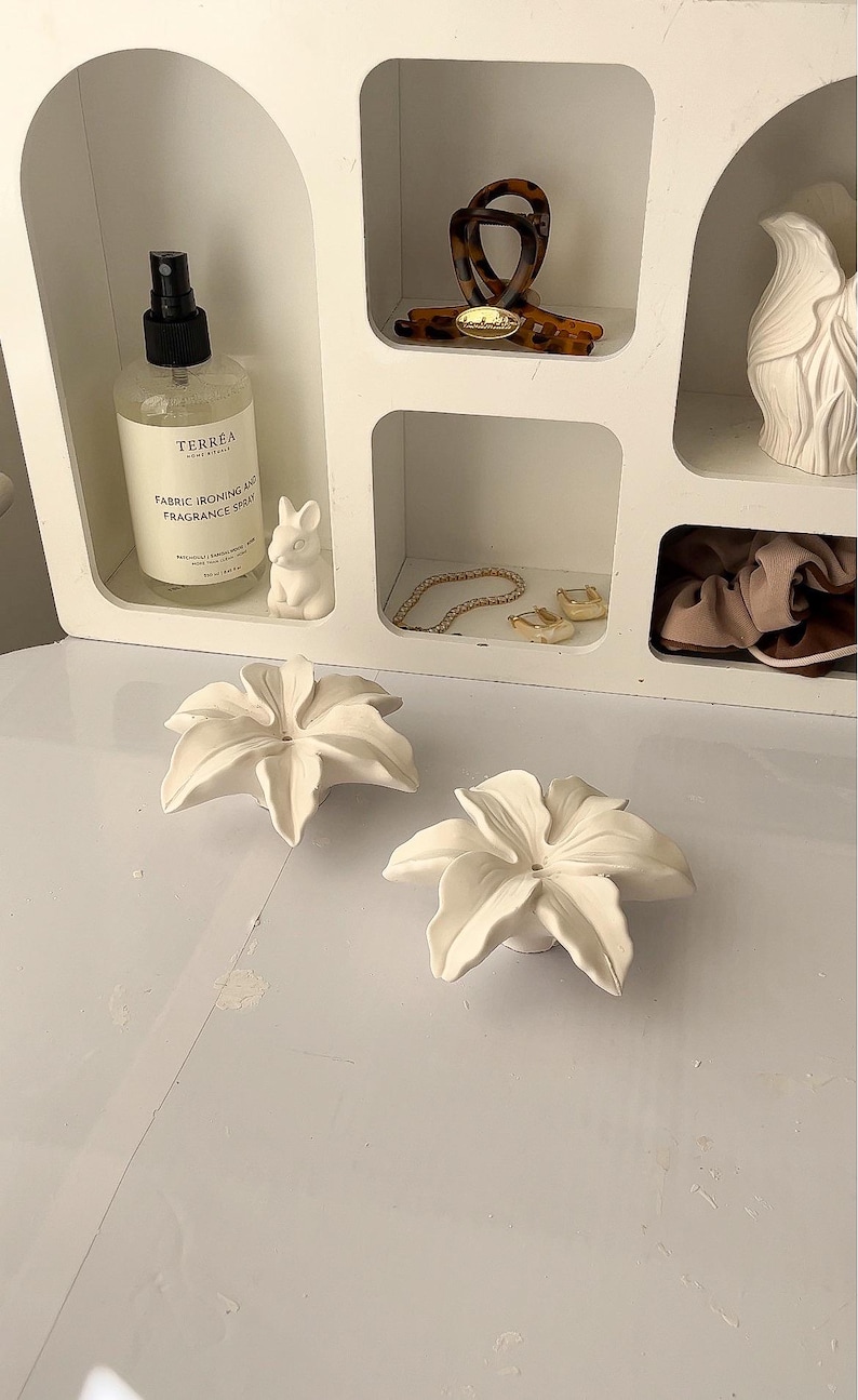 May include: Two white ceramic flower-shaped objects sit in the foreground. Behind them, a white shelving unit contains a clear spray bottle labeled Terrea Fabric Ironing and Fragrance Spray, a tortoiseshell hair clip, a white figurine, and jewelry.