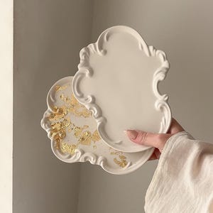 May include: Two off-white, ornate decorative trays. One tray has gold leaf accents. The trays have a scalloped edge and a classic, elegant design. The trays are held by a person.