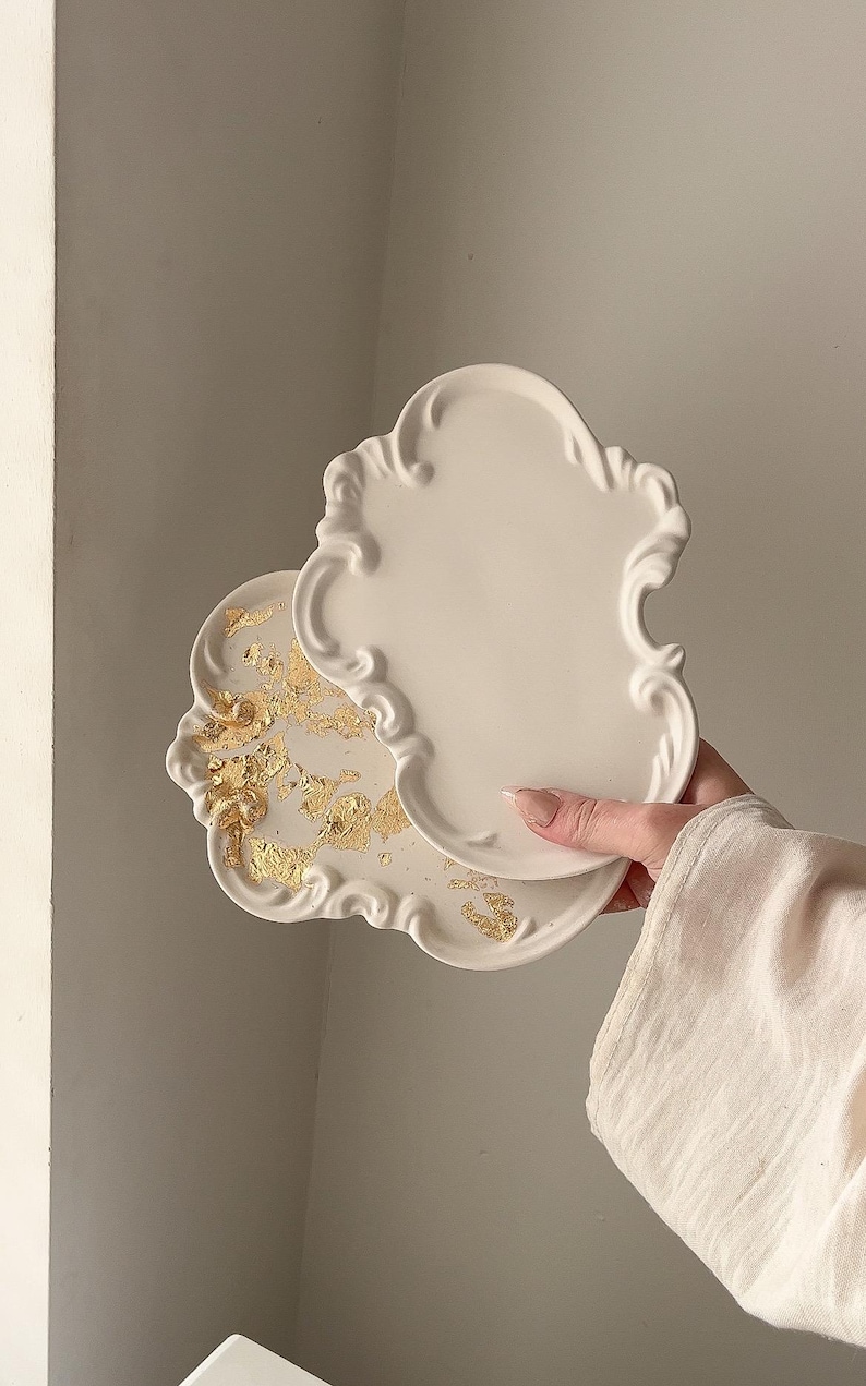 May include: Two off-white, ornate decorative trays. One tray has gold leaf accents. The trays have a scalloped edge and a classic, elegant design. The trays are held by a person.