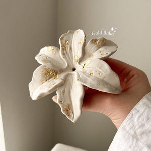 May include: A white ceramic flower sculpture with six petals, featuring a textured surface and scattered gold flake accents. The center of the flower has a small circular opening. The text "Gold flake" is printed in white near the top right.