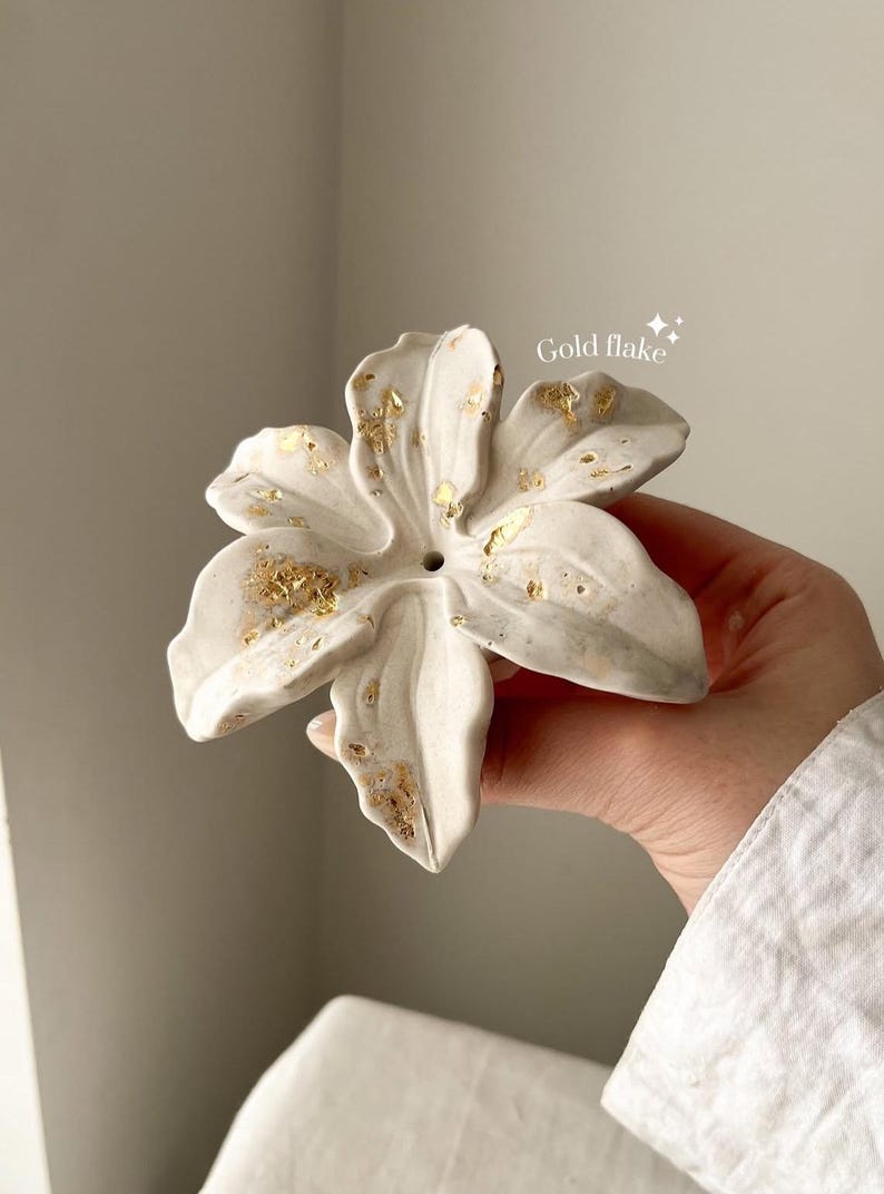 May include: A white ceramic flower sculpture with six petals, featuring a textured surface and scattered gold flake accents. The center of the flower has a small circular opening. The text "Gold flake" is printed in white near the top right.