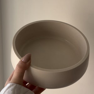 May include: A beige, round bowl with a smooth, matte finish. The bowl has a wide, shallow design with a curved rim. The neutral color and simple shape suggest a modern, minimalist aesthetic. This bowl could be used for serving or as a decorative piece.