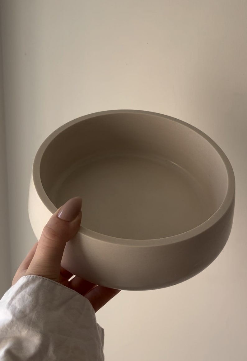May include: A beige, round bowl with a smooth, matte finish. The bowl has a wide, shallow design with a curved rim. The neutral color and simple shape suggest a modern, minimalist aesthetic. This bowl could be used for serving or as a decorative piece.