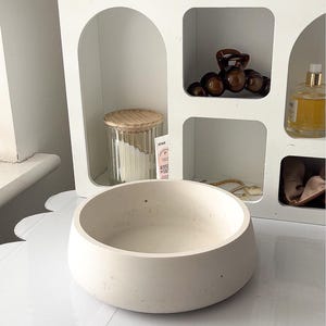 May include: A light beige, round, concrete bowl sits on a white surface. A white shelf with arched and square openings is in the background, holding a glass jar with a wooden lid, hair accessories, and a perfume bottle.