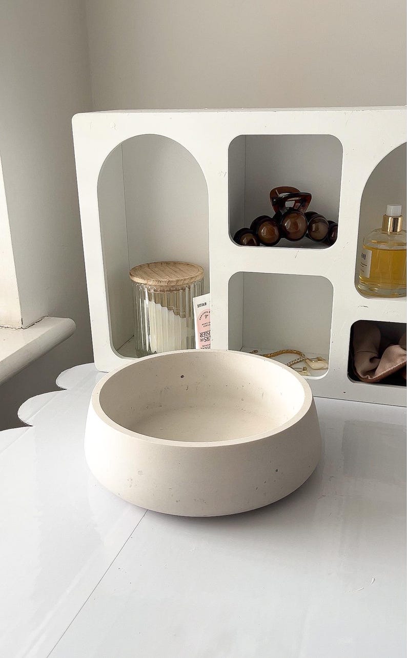 May include: A light beige, round, concrete bowl sits on a white surface. A white shelf with arched and square openings is in the background, holding a glass jar with a wooden lid, hair accessories, and a perfume bottle.