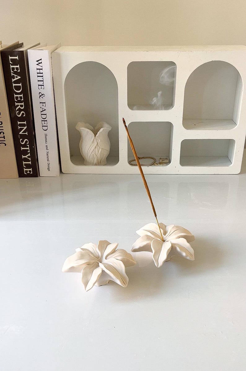 May include: Two off-white ceramic incense holders shaped like blooming flowers. One holder contains a thin, brown incense stick with a wisp of smoke rising from the tip. The holders have layered, curved petals and a matte finish.