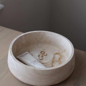 May include: A light-colored wooden bowl with a cream-colored tube of lotion, gold earrings, and a gold beaded necklace. The bowl has a marbled appearance and sits on a light-colored wooden surface.
