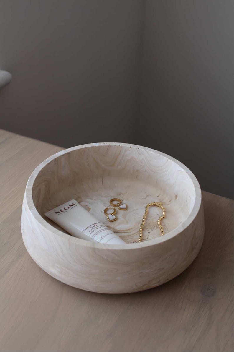 May include: A light-colored wooden bowl with a cream-colored tube of lotion, gold earrings, and a gold beaded necklace. The bowl has a marbled appearance and sits on a light-colored wooden surface.