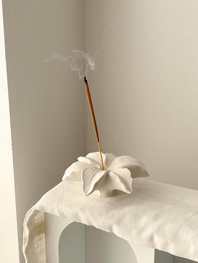 May include: A white ceramic incense holder shaped like a blooming flower with curved petals. A single brown incense stick is inserted into the center of the holder, emitting a thin trail of white smoke that curls upward into the air.