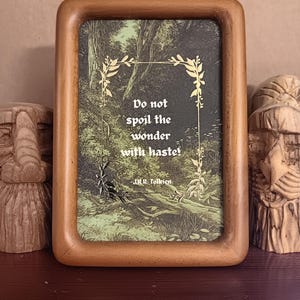 May include: A framed print with a quote from J.R.R. Tolkien, set against a forest scene. The wooden frame surrounds the print, which features the text "Do not spoil the wonder with haste!" in white. Decorative elements frame the text.