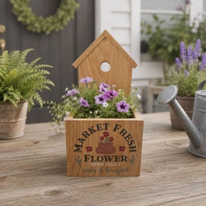 May include: Wooden planter box with a birdhouse top, filled with purple flowers and greenery. The box has the words "MARKET FRESH FLOWER" printed on the front. A watering can and potted plants are in the background.