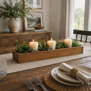 May include: A wooden candle centrepiece featuring three lit pillar candles and greenery. The candles are set in a rectangular wooden box, creating a warm ambiance. The centrepiece is placed on a wooden table with a neutral-coloured runner.