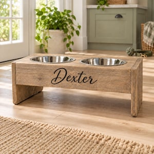 Peut inclure: Un support rectangulaire en bois pour animaux avec deux bols circulaires en acier inoxydable encastrés sur le dessus. L'avant du support en bois brun clair porte le nom Dexter écrit en écriture cursive noire. Le support possède deux pieds latéraux.