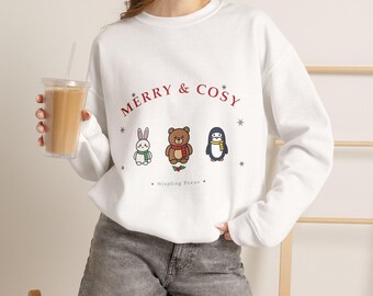 Può includere: Una felpa bianca a girocollo con la scritta rossa MERRY & COSY sopra un grafico raffigurante un coniglio bianco, un orso bruno e un pinguino, ognuno con una sciarpa. Piccoli fiocchi di neve e il testo Wispling Press sono stampati sotto.