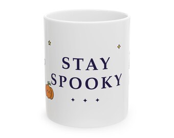 Può includere: Una tazza in ceramica bianca con il testo blu scuro STAY SPOOKY centrato sul davanti. Il design include una piccola illustrazione di una zucca arancione con un volto, insieme a diversi piccoli accenti a forma di stella a quattro punte.
