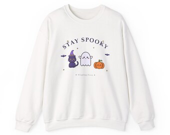 May include: A white long-sleeve sweatshirt featuring the text STAY SPOOKY in dark blue. The graphic includes a black cat wearing a purple witch hat, a white ghost, and an orange pumpkin, all with cute faces, plus small bats and stars.