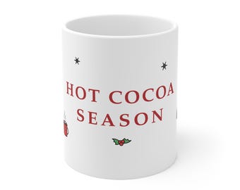 May include: A white ceramic mug featuring the red text HOT COCOA SEASON printed in the centre. The design includes small black star illustrations, a tiny red mug icon with steam, and a small green holly leaf graphic with three red berries.