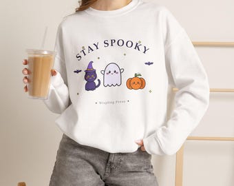 May include: A white long-sleeve sweatshirt featuring the text STAY SPOOKY in dark blue letters. The graphic includes a purple-hatted black cat, a white ghost, and an orange pumpkin, all with smiling faces, plus small bats and stars.