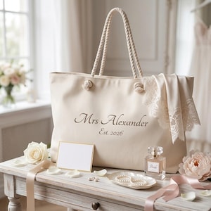 May include: A cream-colored canvas tote bag with rope handles, featuring the dark brown text Mrs Alexander and Est. 2026. A white lace fabric piece is draped over the right side of the bag. The bag is positioned near a perfume bottle and a blank card.