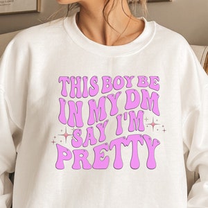 May include: White sweatshirt with the text "THIS BOY BE IN MY DM SAY I'M PRETTY" in pink bubble letters. The design includes star accents. The bottom of the image has the text "GRAPIKA" and "SVG | PNG | JPG".