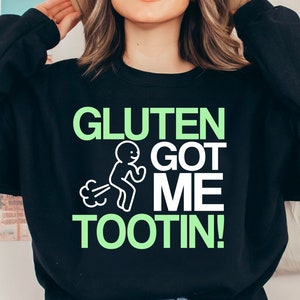 May include: Black sweatshirt with the text "GLUTEN GOT ME TOOTIN!" in green and white. The design includes a stick figure running and emitting gas. The sweatshirt is a casual, humorous apparel item.