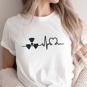 Puede incluir: Camiseta blanca con un gráfico negro de un símbolo de radiación, una línea de latido cardíaco y un corazón. La camiseta tiene mangas cortas y está hecha de un material suave. La palabra "GRAPIKA" está impresa en la esquina inferior derecha.