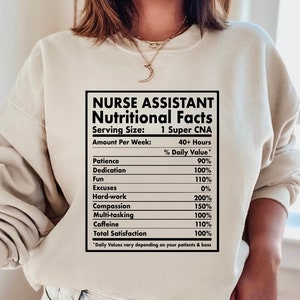 Puede incluir: Una sudadera beige con un gráfico negro que dice "Nurse Assistant Nutritional Facts" con una tabla que enumera el valor diario de varias cualidades necesarias para el trabajo, como la paciencia, la dedicación, la diversión, el trabajo duro, la compasión, la multitarea, la cafeína y la satisfacción total. El gráfico está diseñado para parecerse a una etiqueta de información nutricional.