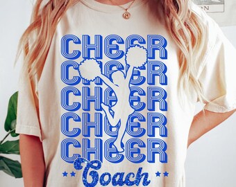 May include: A white t-shirt with a blue and white graphic design of a cheerleader with pom-poms and the text "Cheer Coach" in blue glitter.