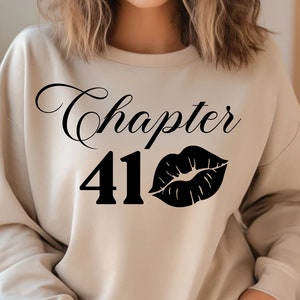 Puede incluir: Una sudadera beige con el texto "Chapter 41" y una impresión de labios negros.